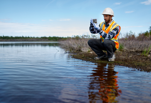 A water scientist kneels down along a river and examines a water sample.